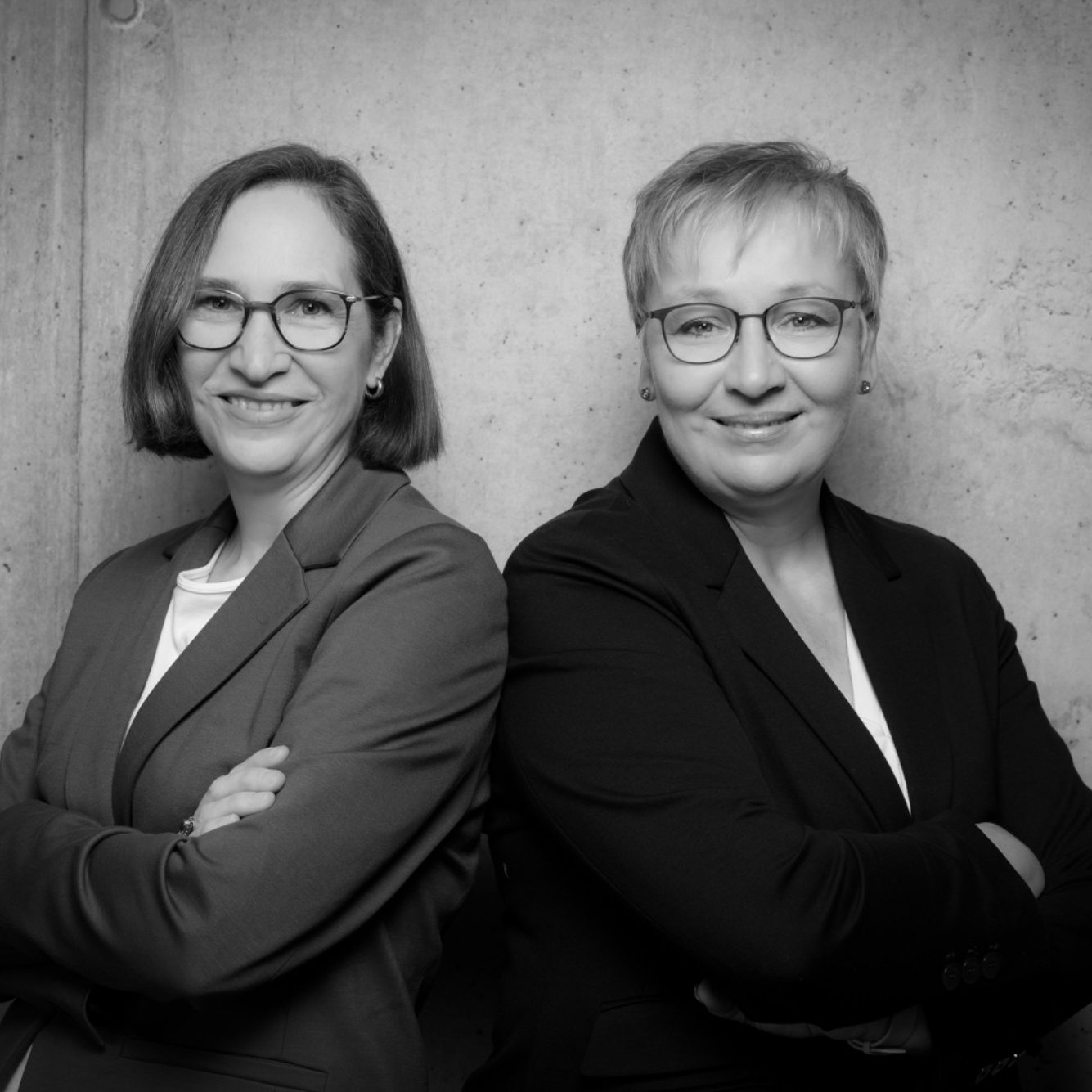 Iris Bothe und Petra Ringmann stehen lächelnd mit verschränkten Armen vor einer grauen Wand.
